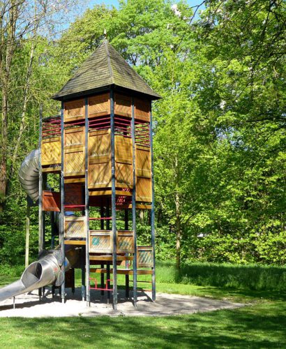Spielplatz aus Holz mit Rutsche. Große Bäume umringen den Spielplatz