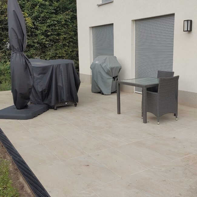 Steinterrasse vor modernen Haus umgeben von einer Grünfläche. Terrasse umrandet von schwarzem Stein