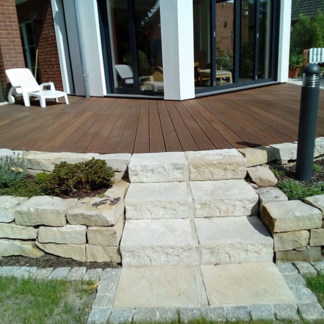 Steintreppe führt zu einer erhöhten Holzterrasse umgeben von Mauern und Beeten mit eine, Liegestuhl